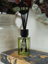 A reed diffuser bottle with pale green liquid and black reeds, labeled &