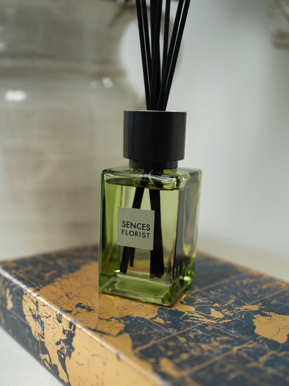 A Sences Florist reed diffuser with a square green glass bottle, black cap, and black reeds, resting on a gold and navy patterned box.
