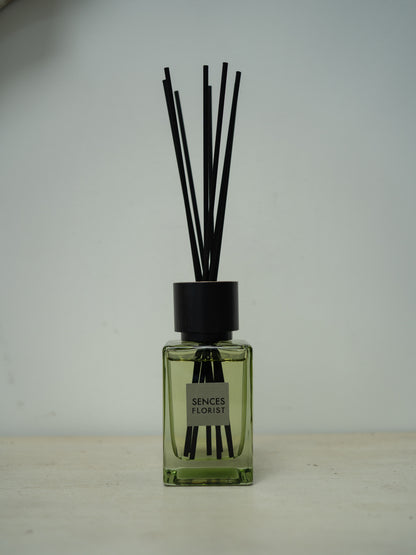 A reed diffuser bottle with a square translucent green glass body, a black cap, and multiple black reeds, with a label reading &