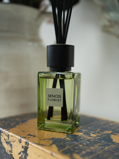 A glass reed diffuser bottle filled with light green liquid, with black reeds and a black cap, labeled &