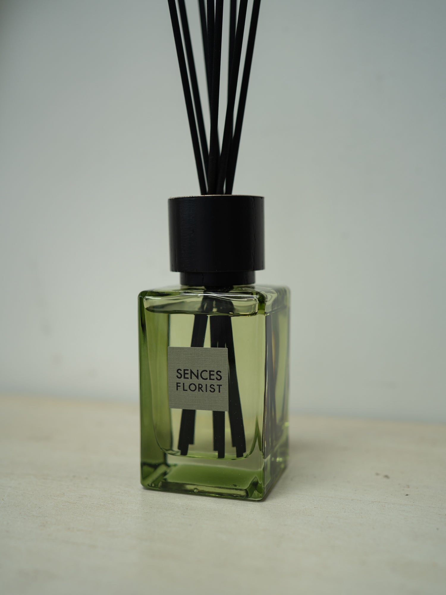 A reed diffuser bottle with a translucent olive-green glass body, a matte black cap, and black reeds, featuring a label that reads &