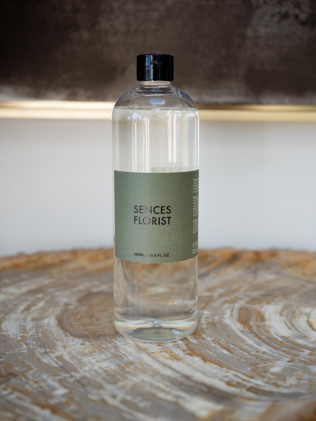A clear glass bottle of Scentes Florist refill bottle with a black cap and a muted sage green label, resting on a rustic wood slice surface.