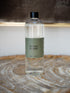A clear glass bottle of Scentes Florist refill bottle with a black cap and a muted sage green label, resting on a rustic wood slice surface.