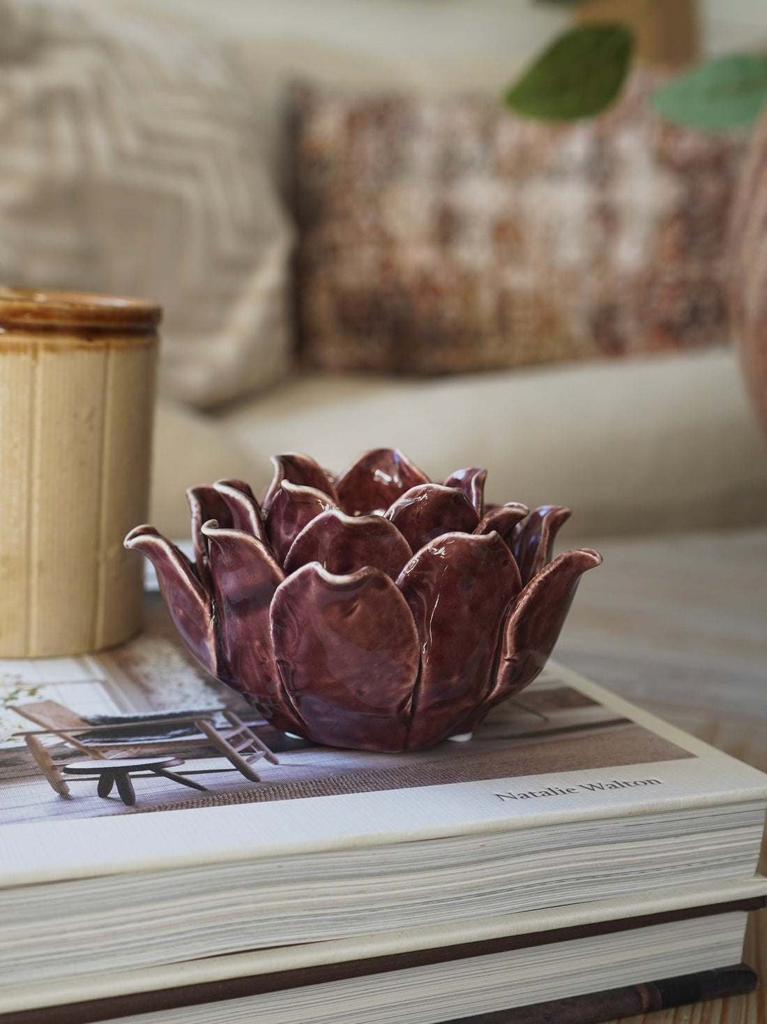 Decorative plum lotus-shaped candle holder on a book with a blurred sofa and plant in the background