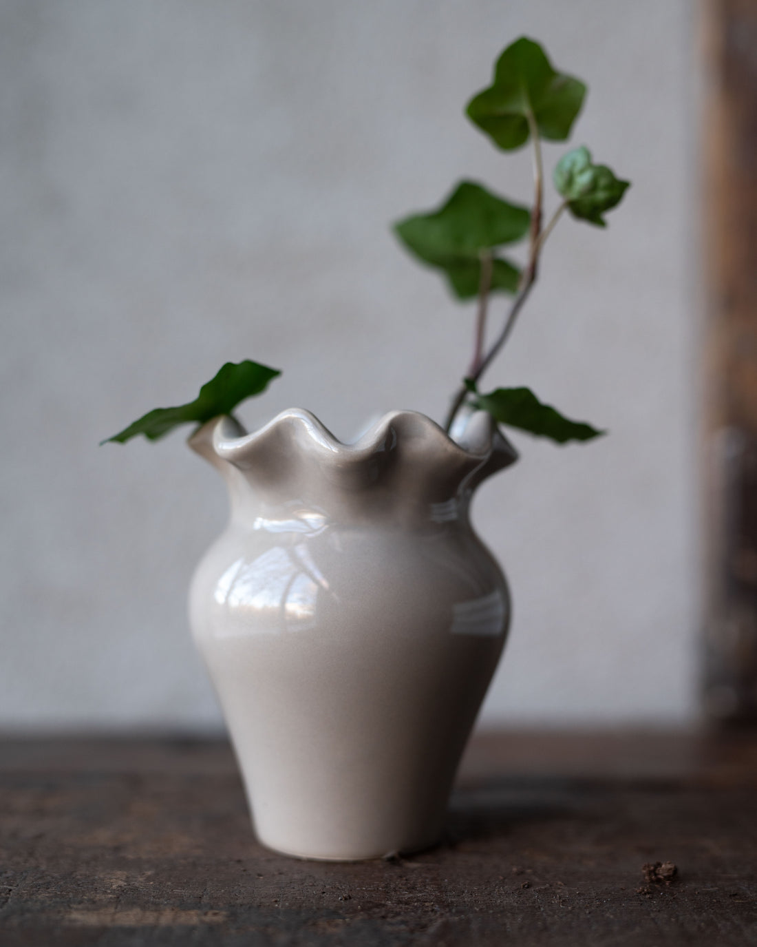 White ceramic vase with a textured surface and ruffled rim, containing a branch with green leaves on a wooden surface.