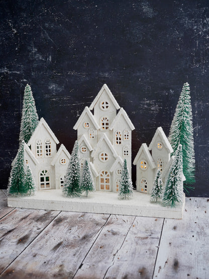 Decorative model of a snowy village with houses and trees on a wooden surface against a dark background.