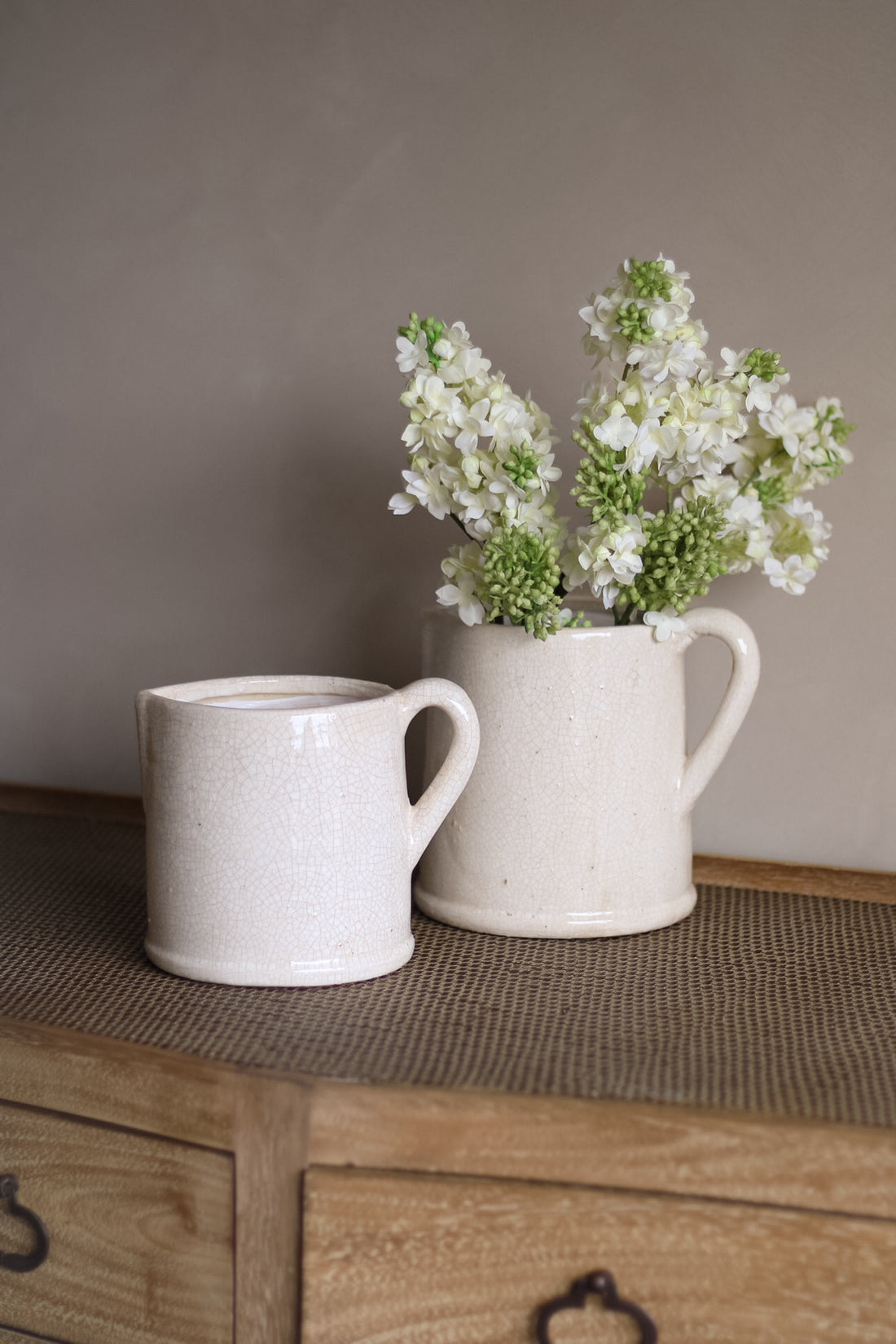 Two ceramic jugs on a wooden surface with one containing white flowers.