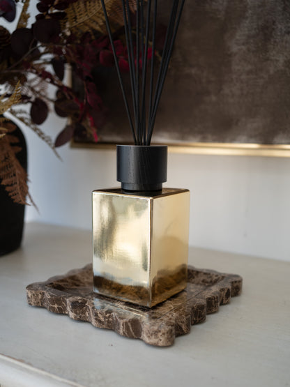 A gold-toned square reed diffuser bottle with a black cap and black reeds, placed on a textured brown stone coaster on a light tabletop.