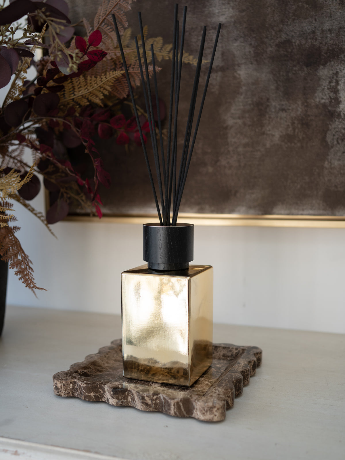 A reed diffuser with a reflective gold square glass bottle, a matte black cap, and black reed sticks, displayed on a rustic stone coaster on a white shelf.
