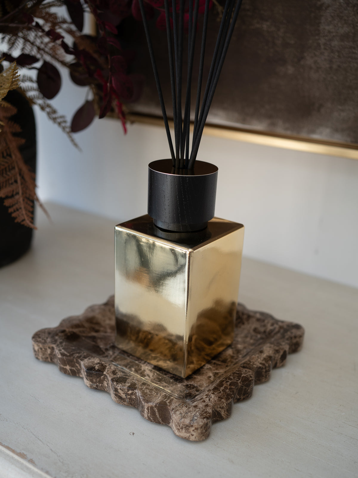 A reed diffuser with a reflective gold cube-shaped base, a black reed holder, and black reeds, displayed on a textured brown stone coaster on a light-colored tabletop.