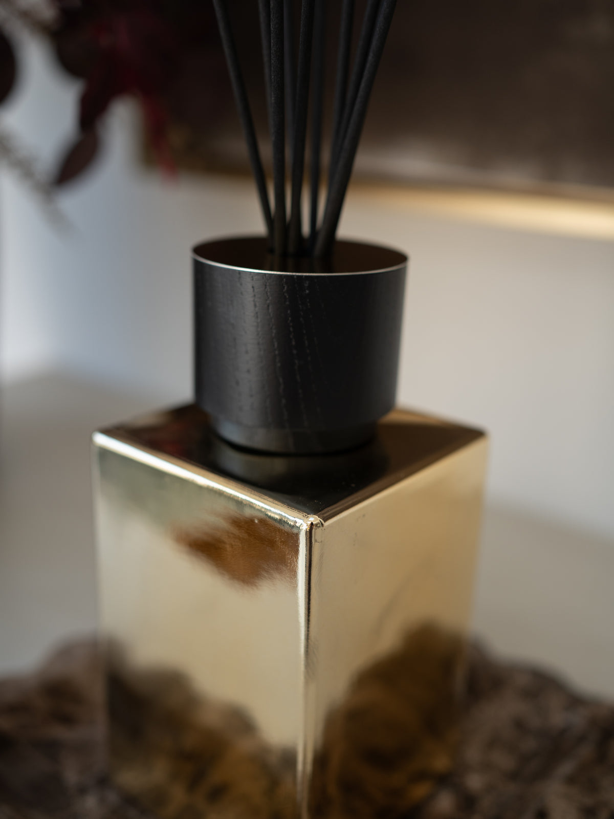 A close-up of a reed diffuser with black reeds in a matte black cylinder, set on a reflective gold cube base against a softly blurred indoor background.