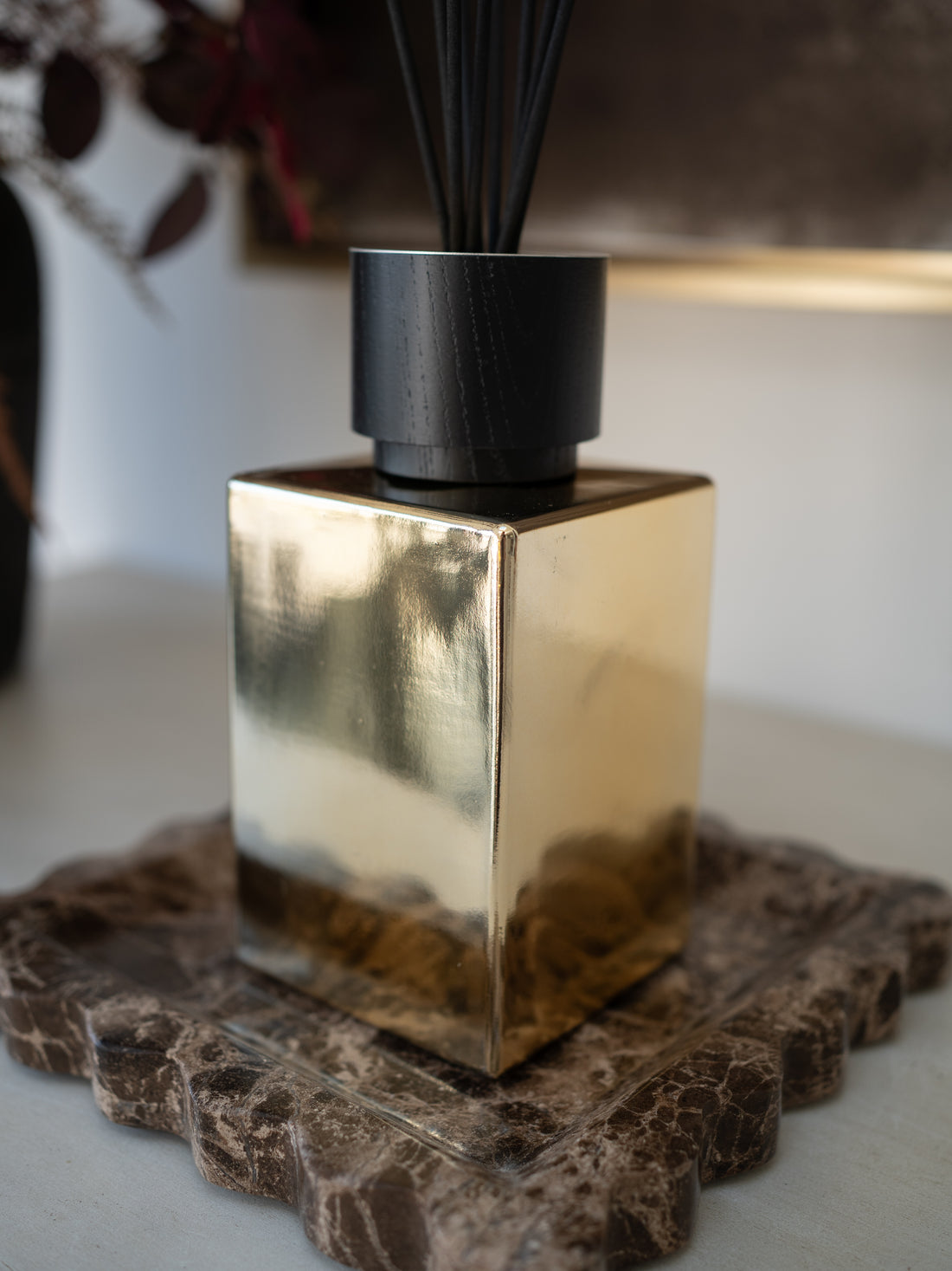 A modern reed diffuser with a reflective gold cube-shaped glass bottle, a matte black cylindrical top, and dark reeds, resting on a brown marble coaster.