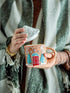 A person wrapped in a green and white striped knit blanket holds a gingerbread house-themed ceramic mug while lifting its white lid. The mug features a peach-brown body with a red window, blue Christmas trees, and white snowflake details.