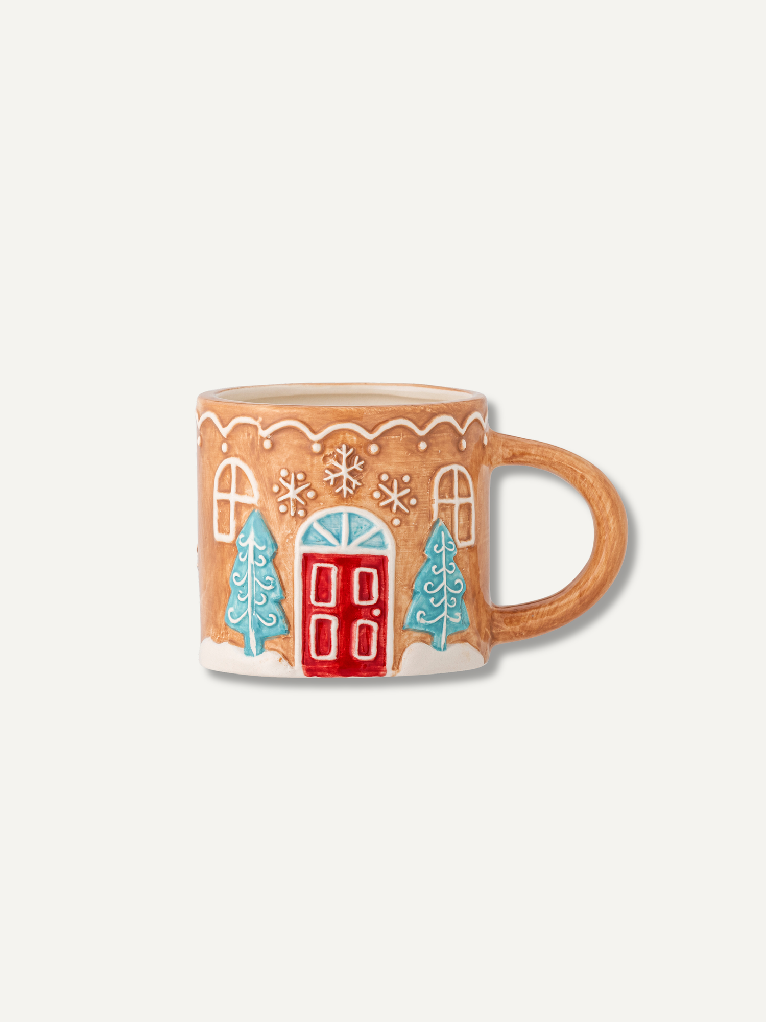 A ceramic mug shaped like a gingerbread house, featuring a warm tan body with white icing-style trim, a red door, and light blue Christmas trees, shown on a plain light gray background.