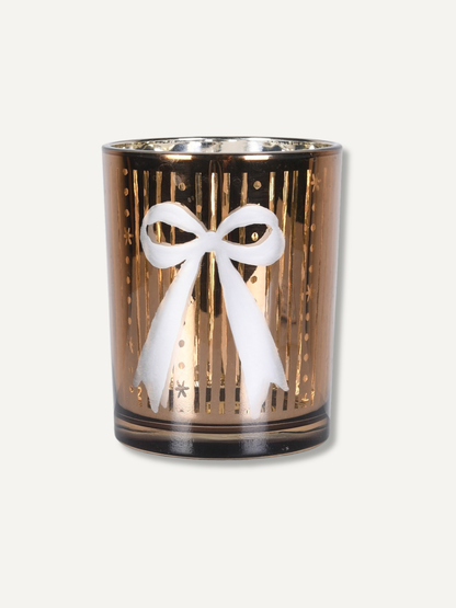 A gold-toned glass candle holder with a white bow design on the front, set against a plain off-white background.