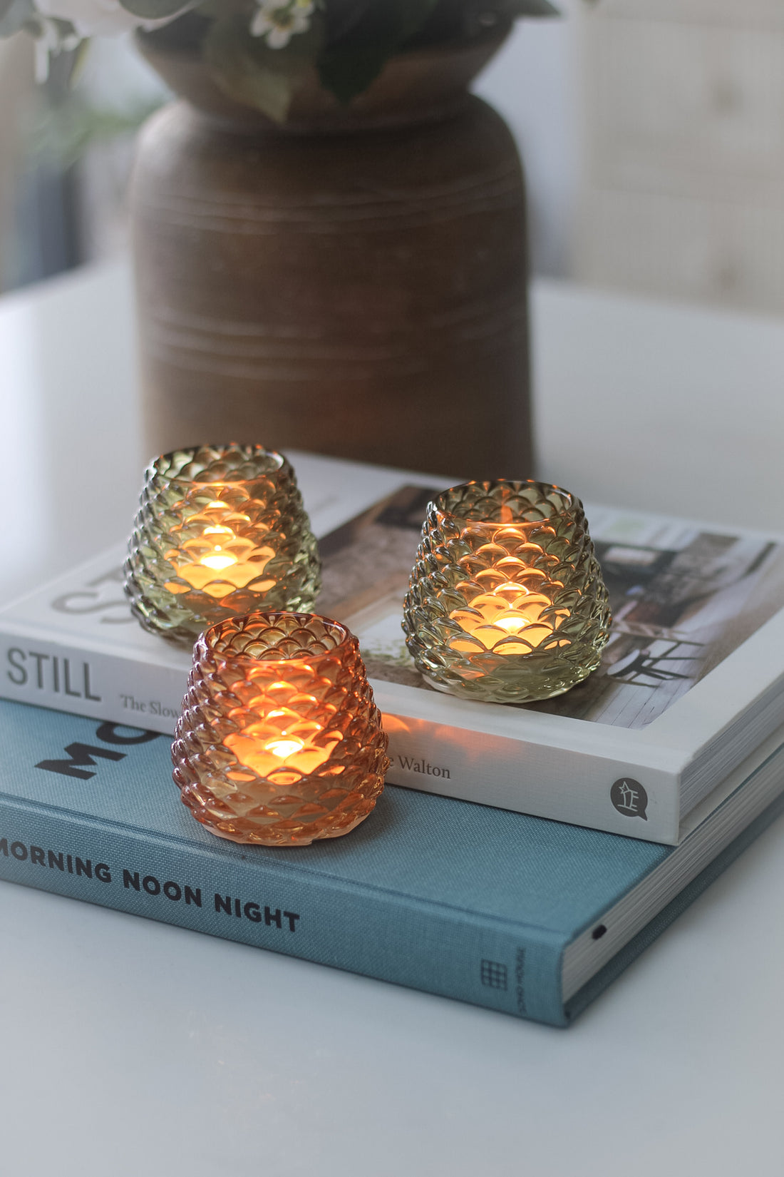 Three glass pinecone votives on a table, with one amber votive lit, placed on top of an open book.