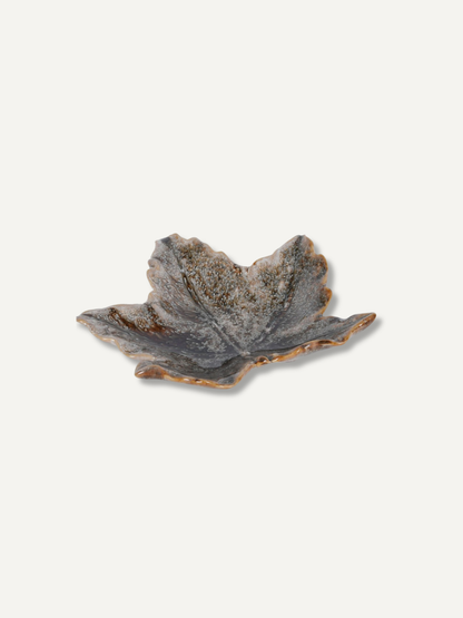 A small leaf-shaped ceramic dish with a textured, speckled glaze in earthy gray, brown, and beige tones, set against a solid off-white background.