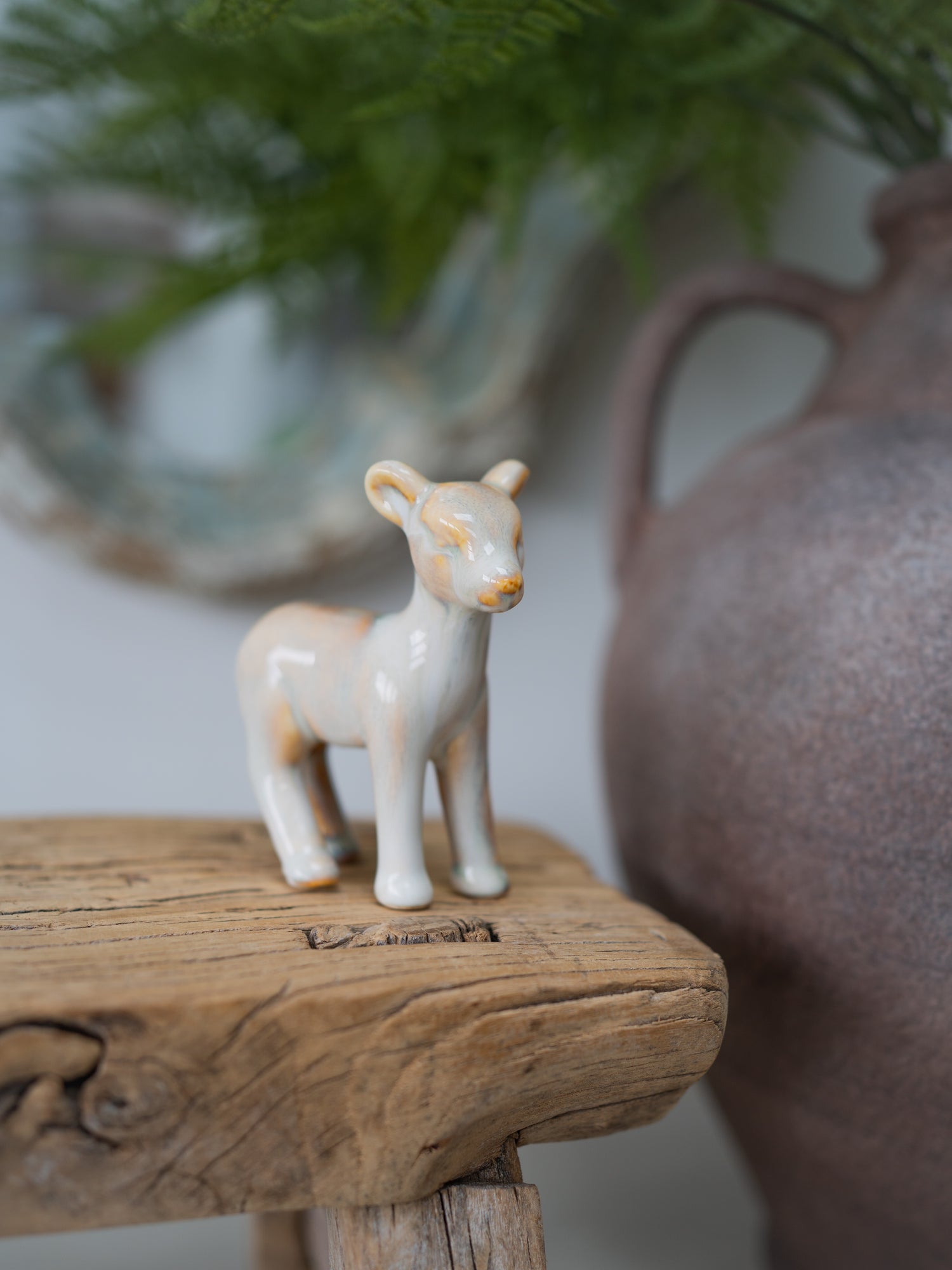 A small ceramic figurine of a standing deer in cream with light tan and muted orange accents, displayed on a rustic wooden surface with a blurred ceramic pitcher and greenery in the background.