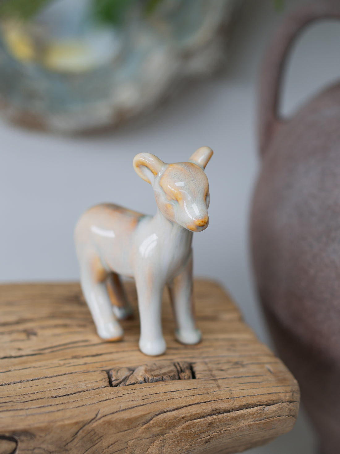 A small ceramic deer with a glossy off-white glaze and subtle warm tan and orange-brown patches, standing on a rustic wooden block with a softly blurred background.