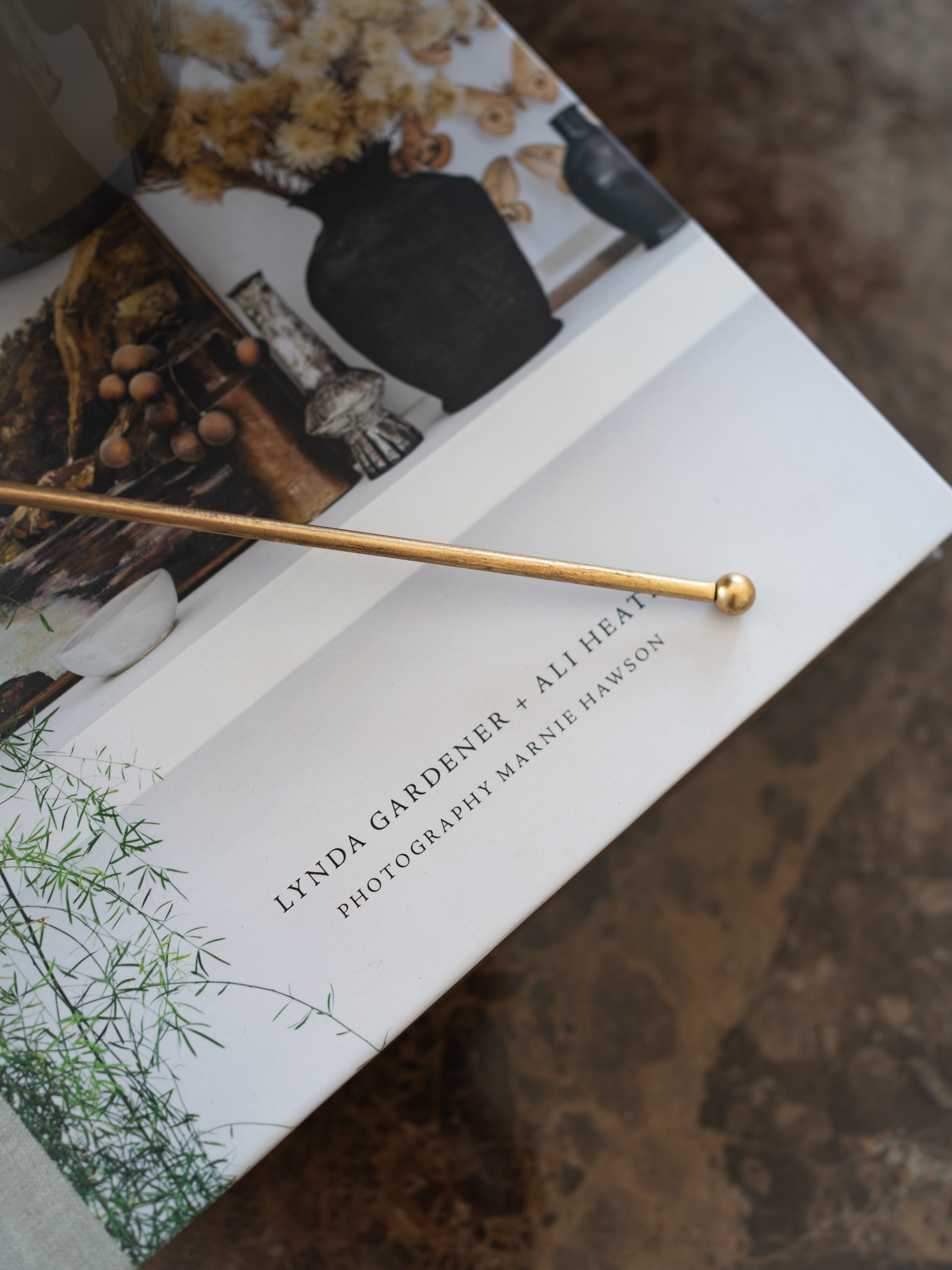 A long, slender gold metal rod with a rounded ball finial lies diagonally across a printed book or magazine cover. The cover shows botanical photography