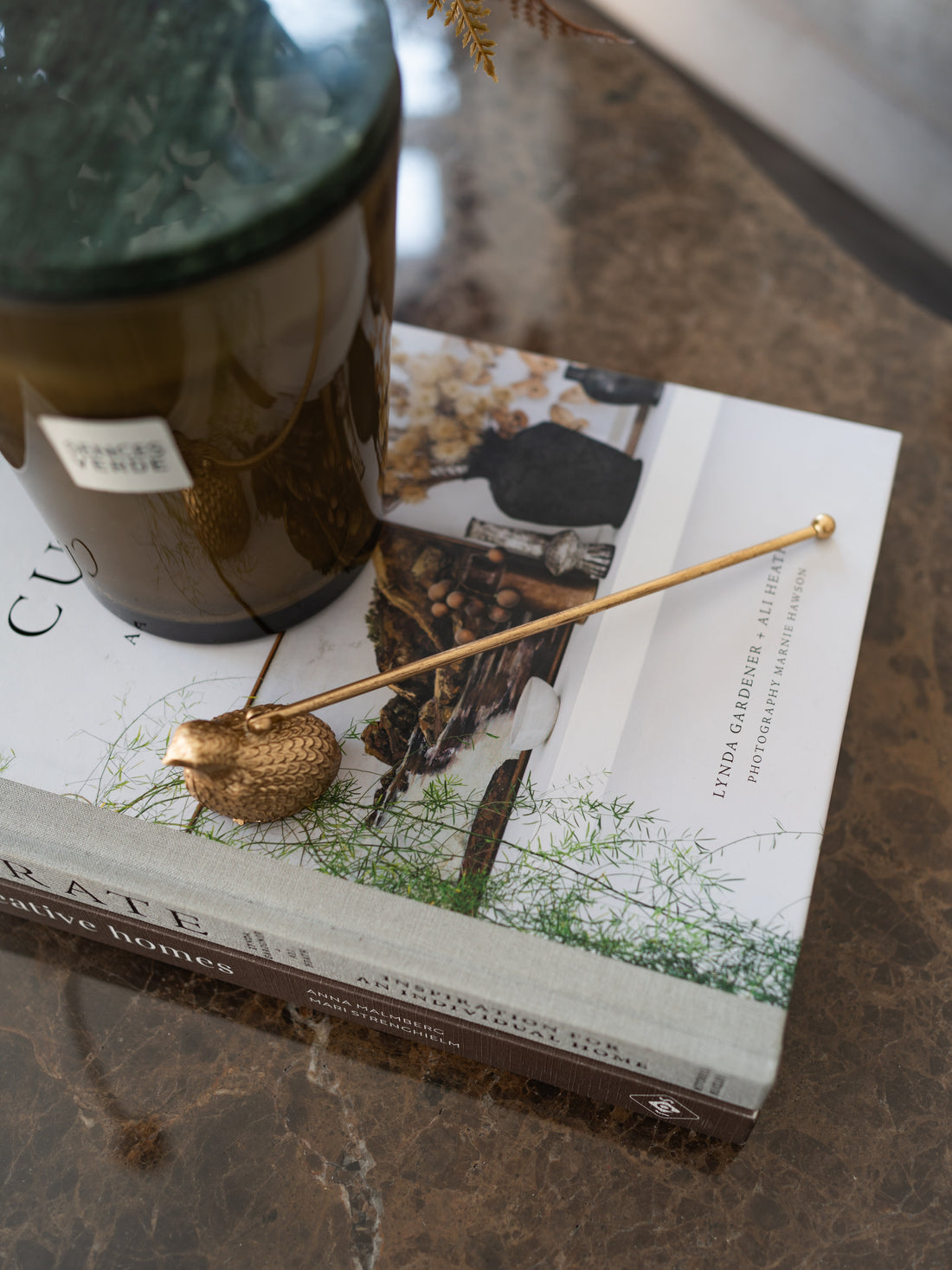 A gold metal bird-shaped candle snuffer with a long straight needle tail rests on a white book with botanical illustrations, next to a dark amber glass jar, all placed on a brown marble surface.