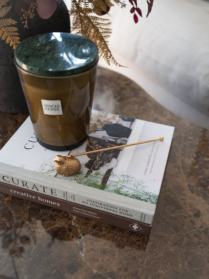 A candle in a dark amber glass jar with a green marbled lid sits on top of a book on a polished marble surface. A small decorative dried seed pod and thin rod rest on the book, with another book underneath and a white fabric background in the upper right.