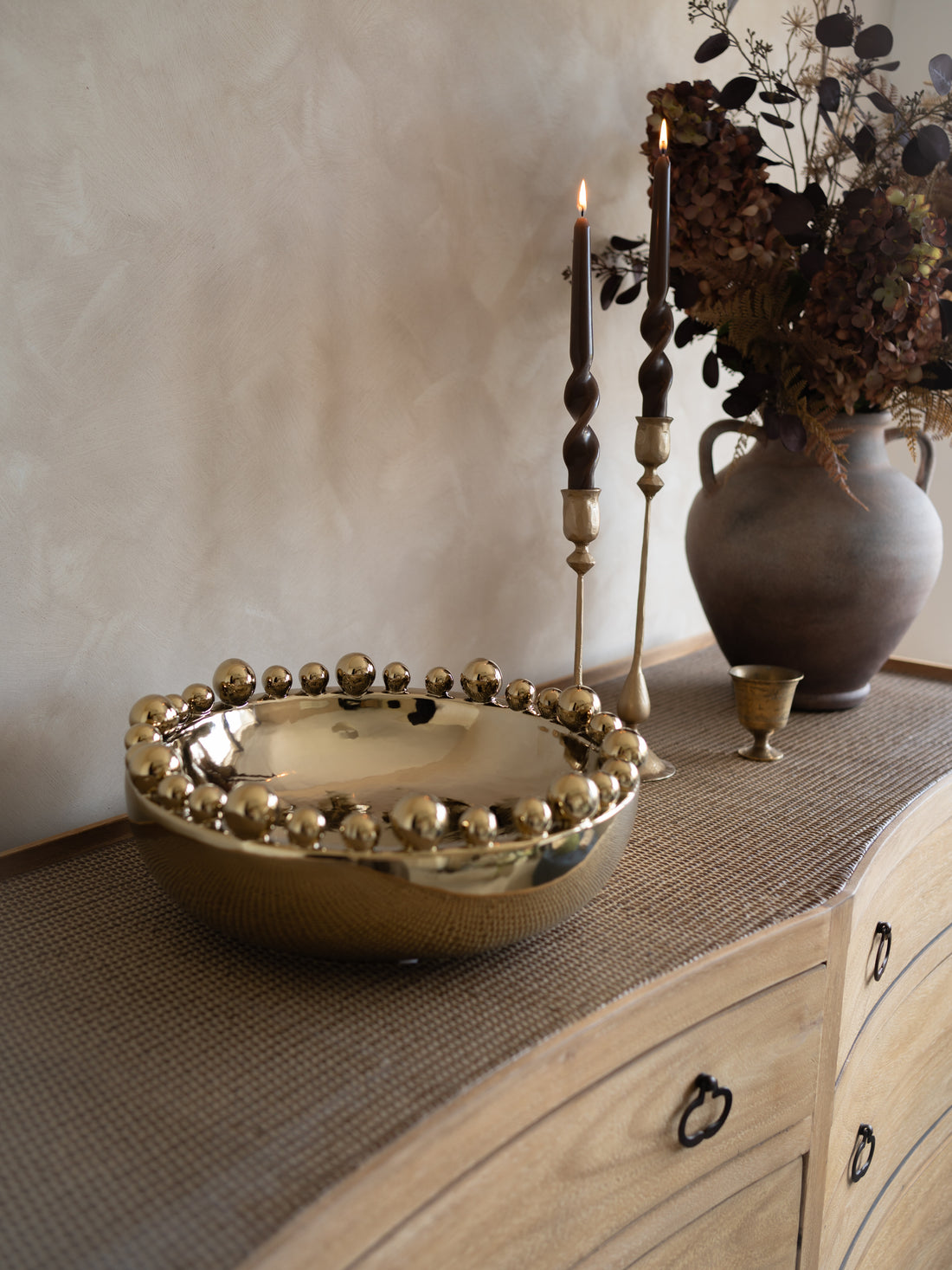 Decorative gold bowl on a wooden surface with candles and a vase in the background.