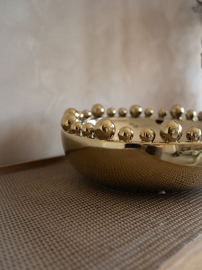 Gold decorative bowl with bobble embellishments on a textured surface.