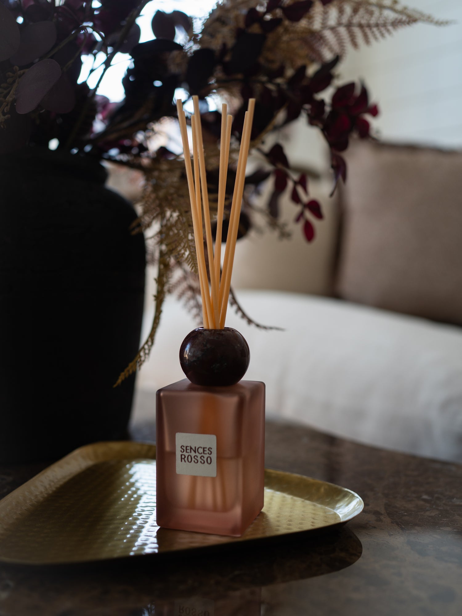 A Scentes Rosso reed diffuser with a , dark burgundy round marble stopper, and natural wood reeds, displayed on a gold tray in a styled home setting.