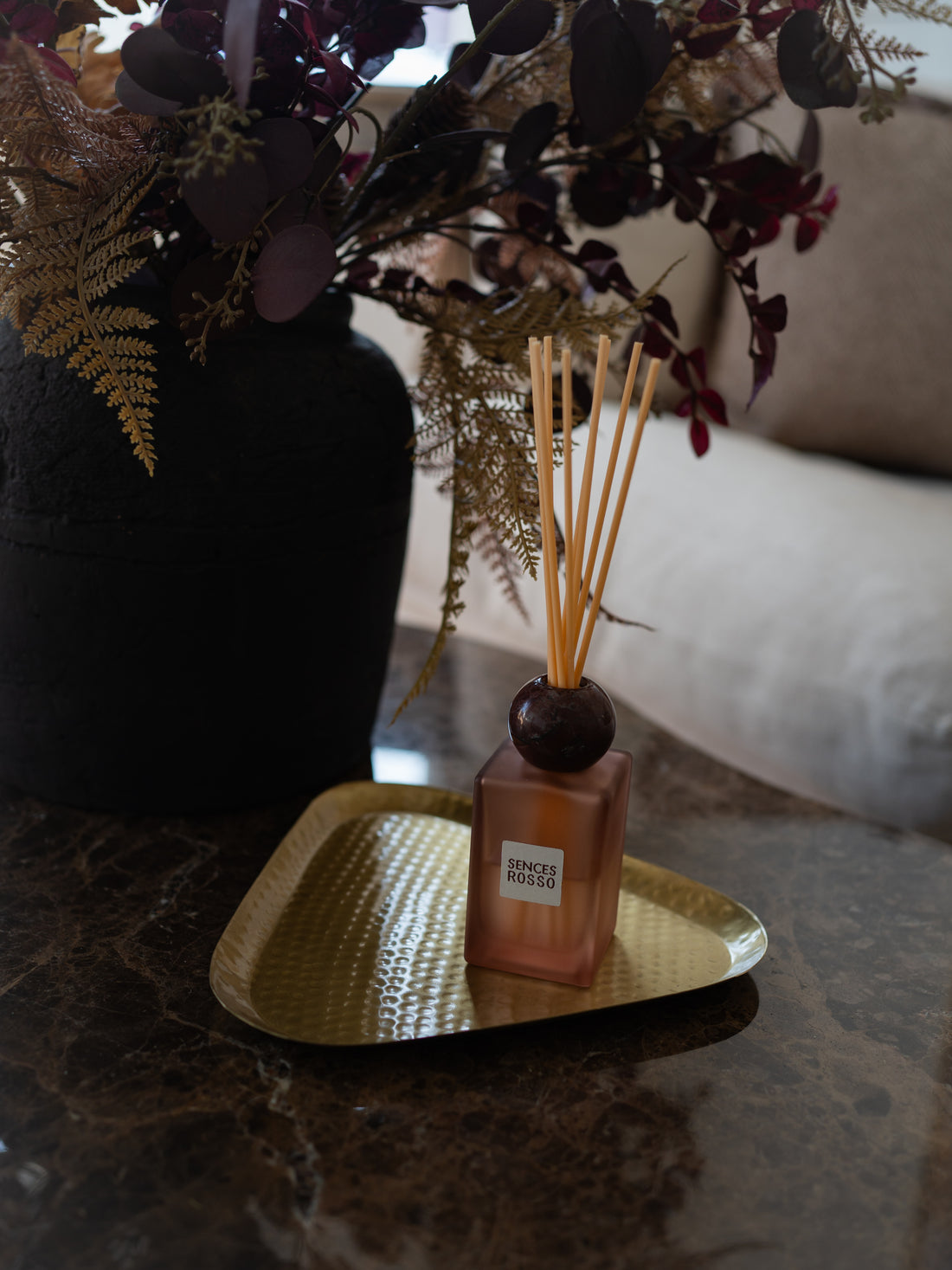 A reed diffuser bottle with a square amber-brown glass body, dark round stopper, and natural reed sticks, resting on a gold tray on a dark marble surface. A large black vase with dried foliage sits behind it in a softly lit interior setting.