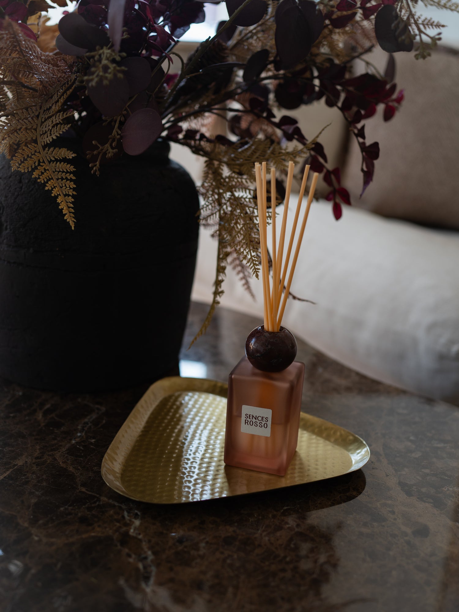 A reed diffuser bottle with a square amber-brown glass body, dark round stopper, and natural reed sticks, resting on a gold tray on a dark marble surface. A large black vase with dried foliage sits behind it in a softly lit interior setting.