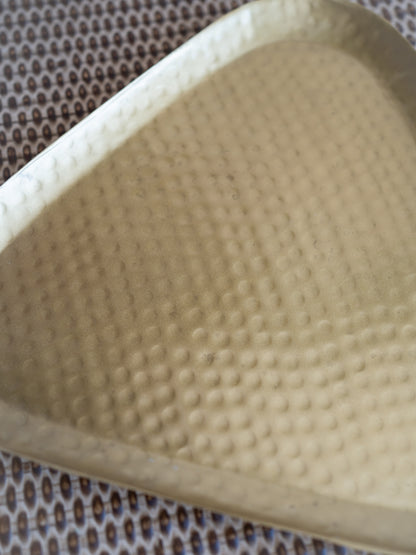 A close-up of a triangular textured tray or dish with a hammered surface, shown resting on a patterned brown and white mat.