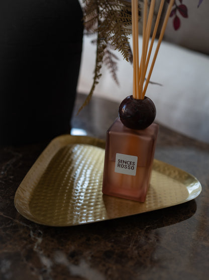 A Senses Rosso reed diffuser bottle with a frosted pinkish-amber glass body, dark wooden stopper, and natural reeds, displayed on a hammered gold tray on a dark marble surface.