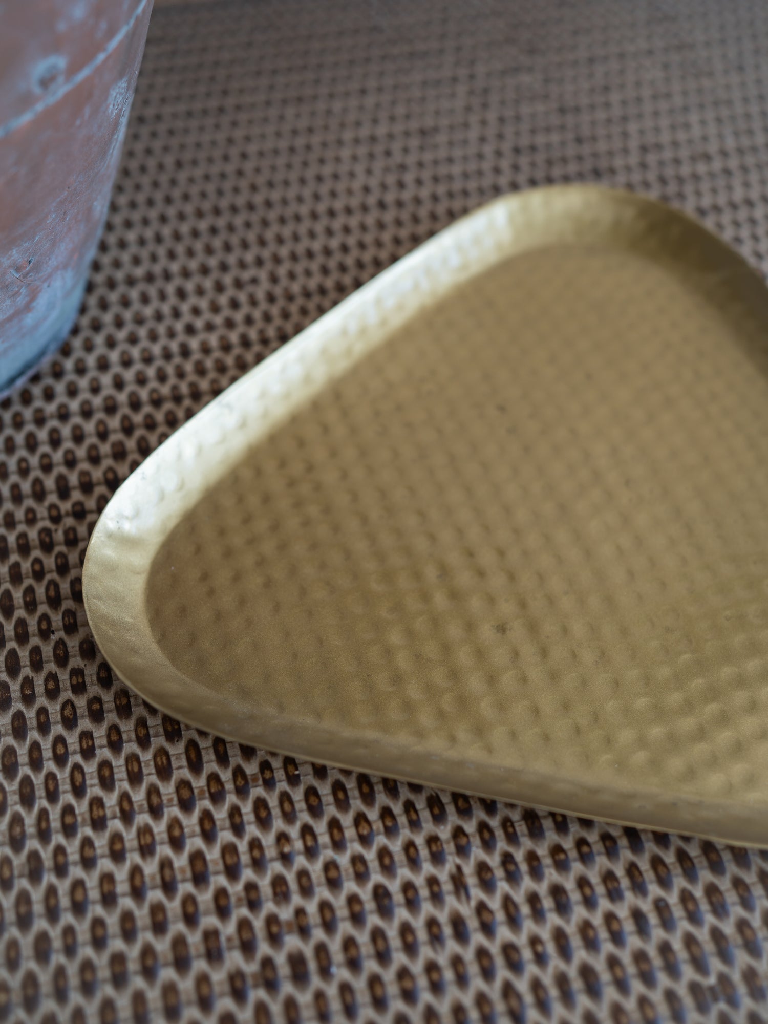 A triangular hammered metal tray with a warm gold finish, resting on a brown textured woven mat.