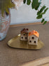 Two miniature ceramic house figurines with one brown roof and one orange roof, displayed on a small gold tray on a woven brown mat.