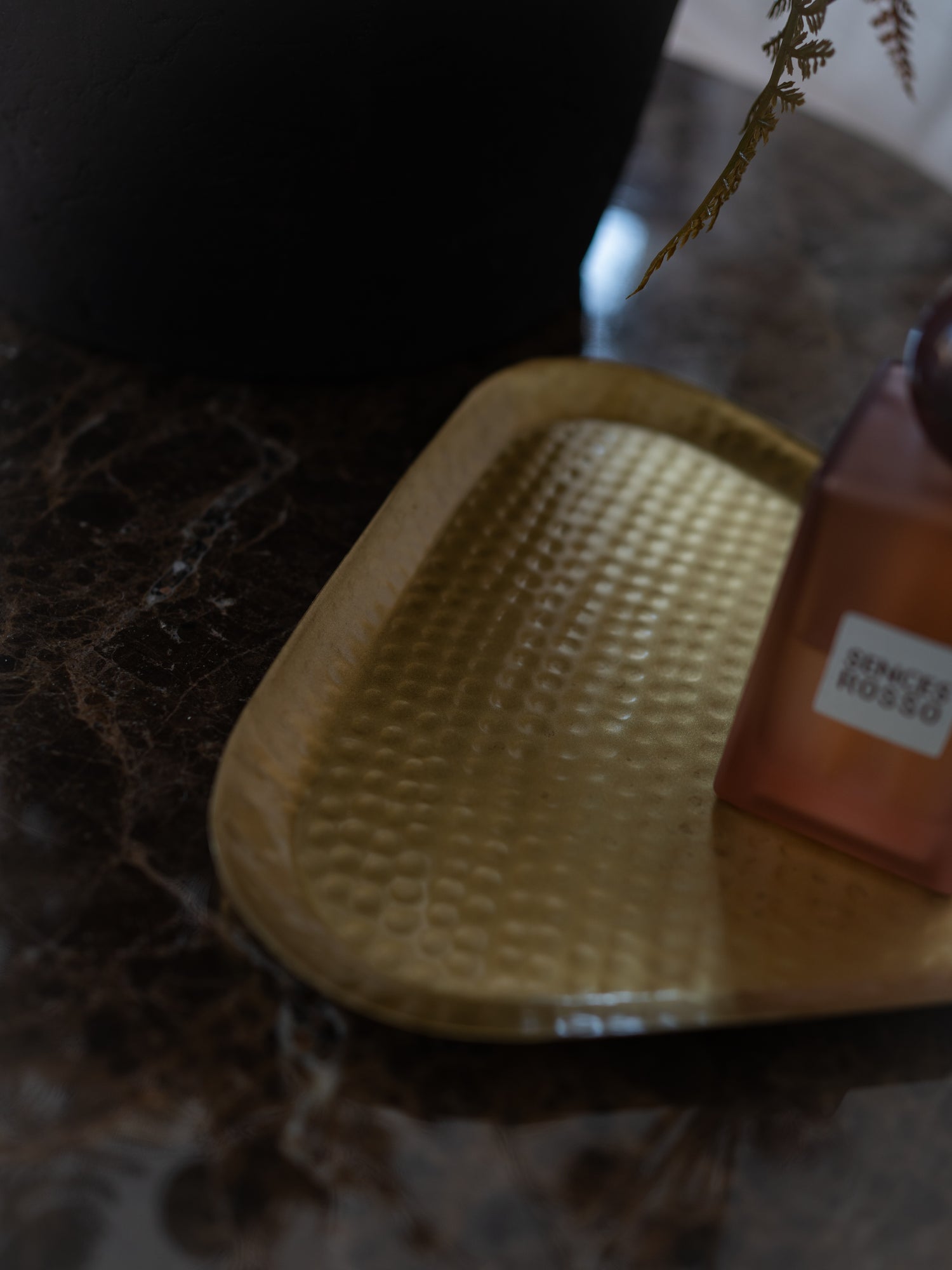 A hammered gold metal tray resting on a dark marble surface next to a brown glass bottle with a white label.