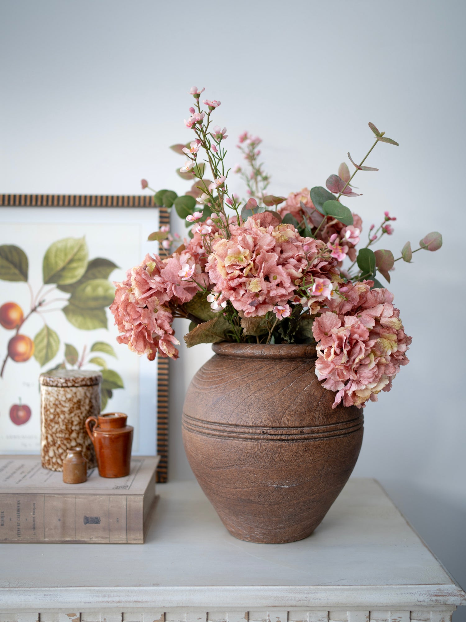 Decorative arrangement of pink flowers in a terracotta vase on a white surface with a light gray wall in the background.