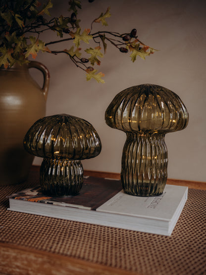 Two ribbed glass mushroom-shaped tabletop lamps pieces in smoky amber-brown tones, displayed on a stack of books on a woven surface with a vase and dried leaves in the background.