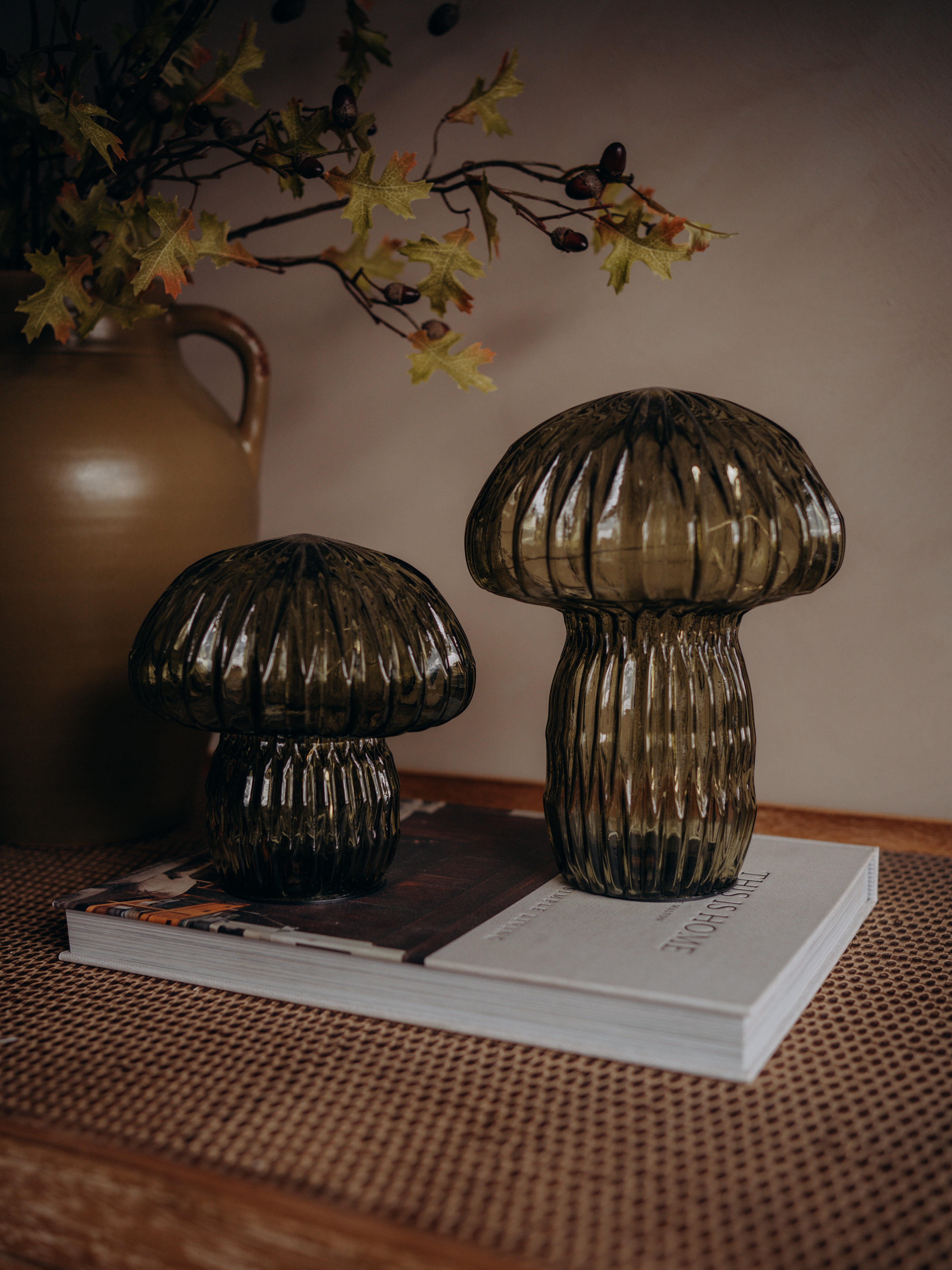 Two ribbed mushroom-shaped glass lamps in dark smoky green, displayed on a stack of books on a woven surface with a ceramic vase and dried leaves in the background.