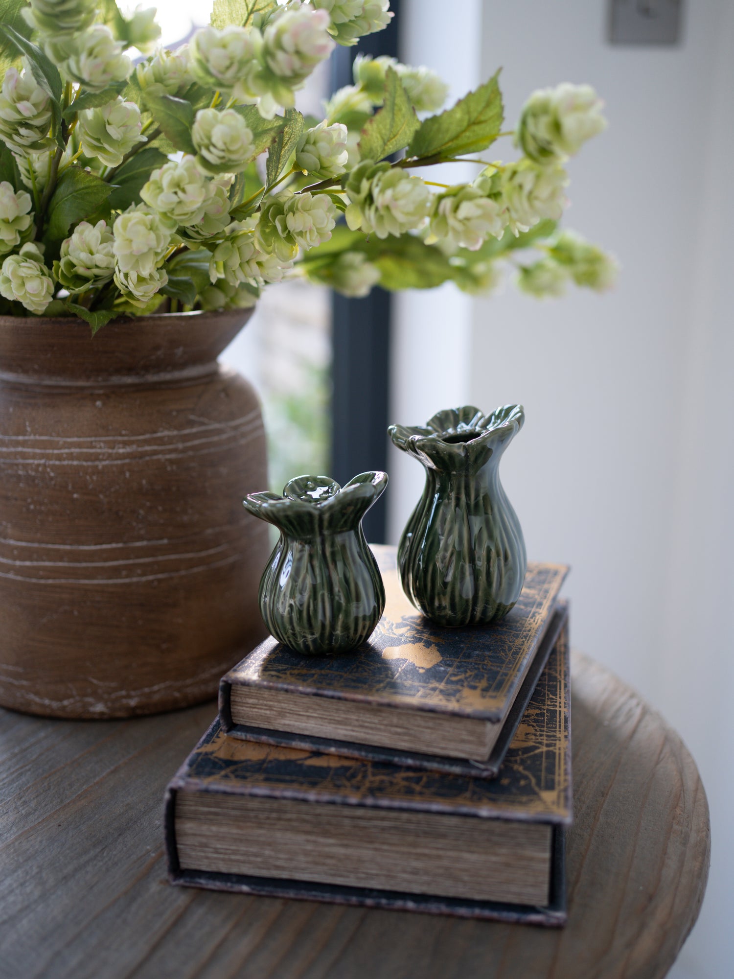 Decorative setup with a large pot of flowers, two small green vases on books, and a blurred background.