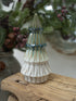 A small ceramic Christmas tree ornament with a ribbed texture, featuring a white base and a blue-green glazed top, displayed on a rustic wooden surface with a blurred holiday background.