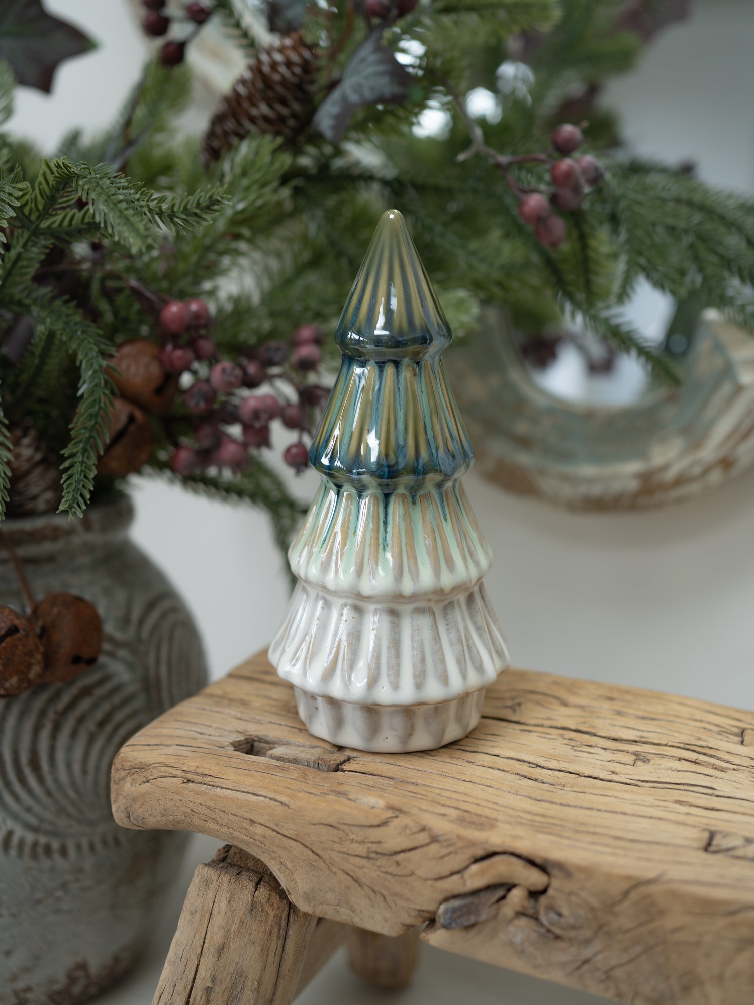 A small ceramic Christmas tree ornament with a ribbed white body and blue-green glazed stripes, displayed on a rustic wooden stool with holiday greenery in the background.