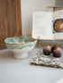 Green ombre ceramic bowl on a white surface with faux figs and a book in the background for kitchen. home styling