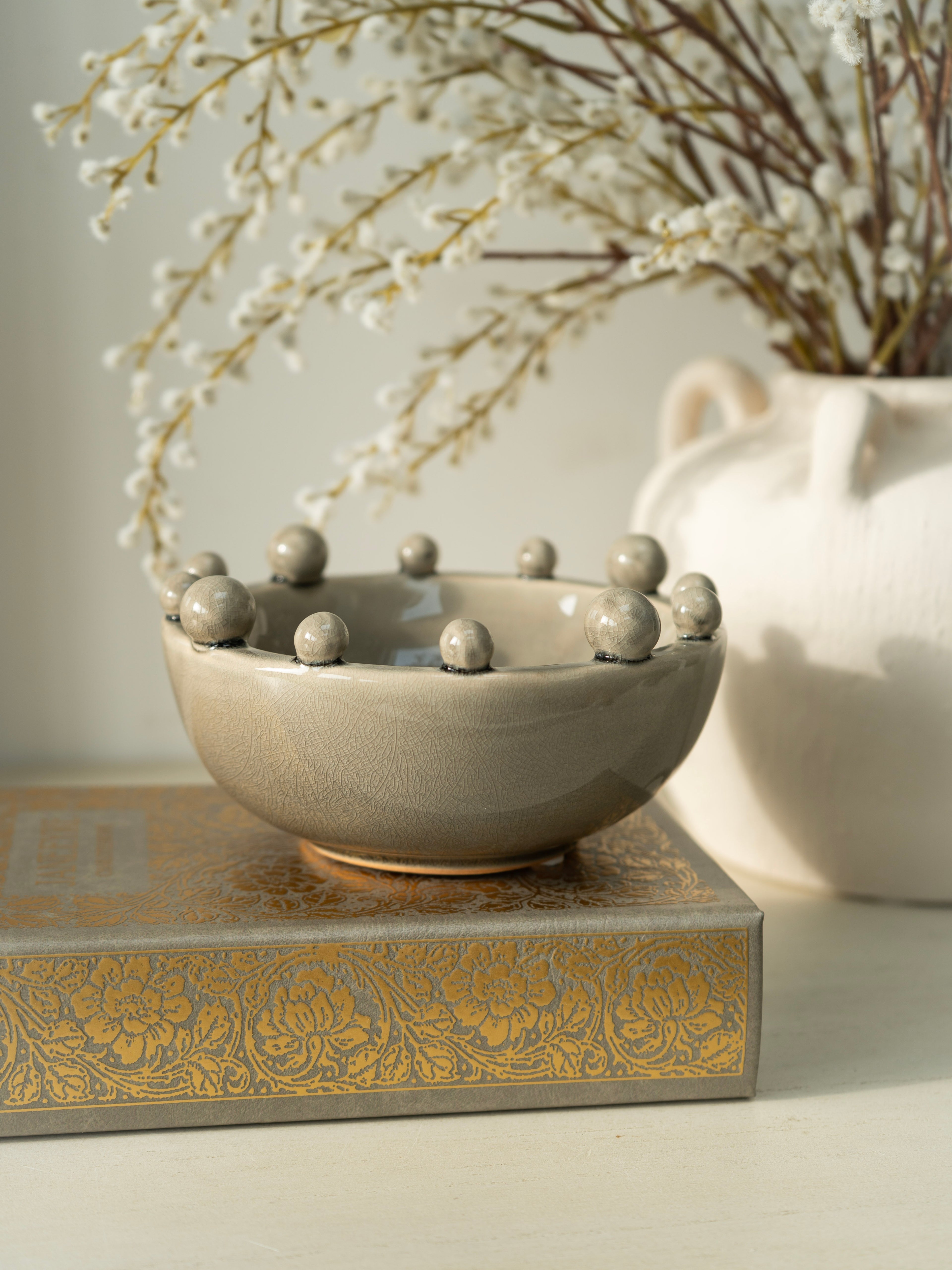 Decorative ceramic bowl with spherical elements on a decorative box with floral patterns, against a neutral background.