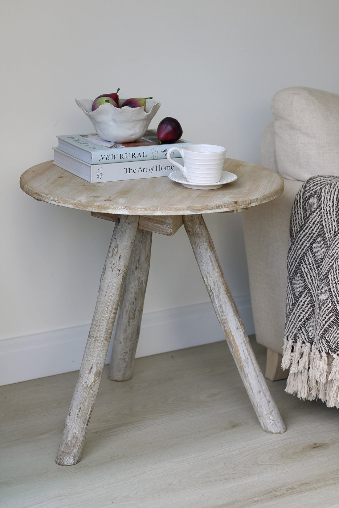 A grey washed round wooden tripod side table with a round top, placed on a hardwood floor next to a couch, with a cup, an apple, and a book on top of it.