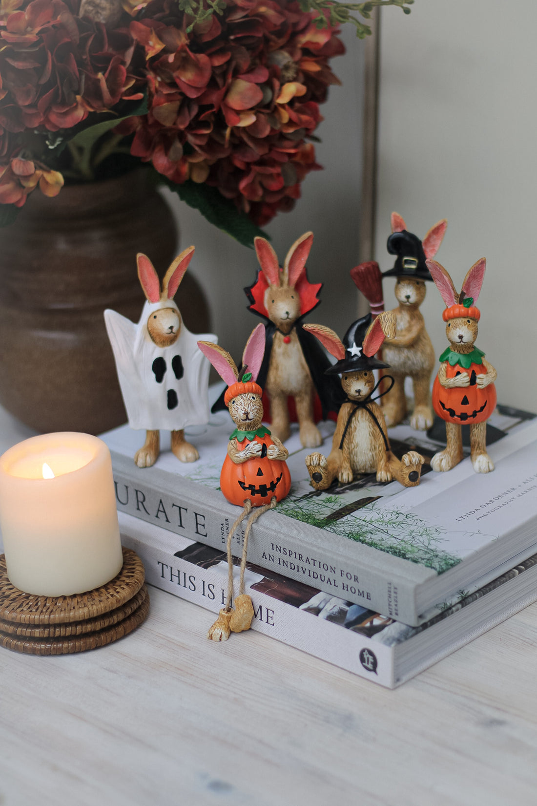 A collection of detailed resin rabbit ornaments with various Halloween-themed designs, such as wearing pumpkin and witch costumes, displayed on a table with a candle and flowers in the background.