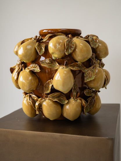 A decorative ceramic vase sculpted with glossy glazed pear and leaves, displayed on a dark pedestal against a light gray background.