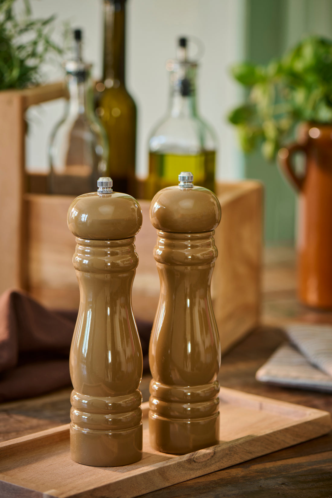 Two brown ceramic pepper and salt mills with silver metal tops, standing on a wooden board in a kitchen setting with olive oil bottles and herbs in the background.