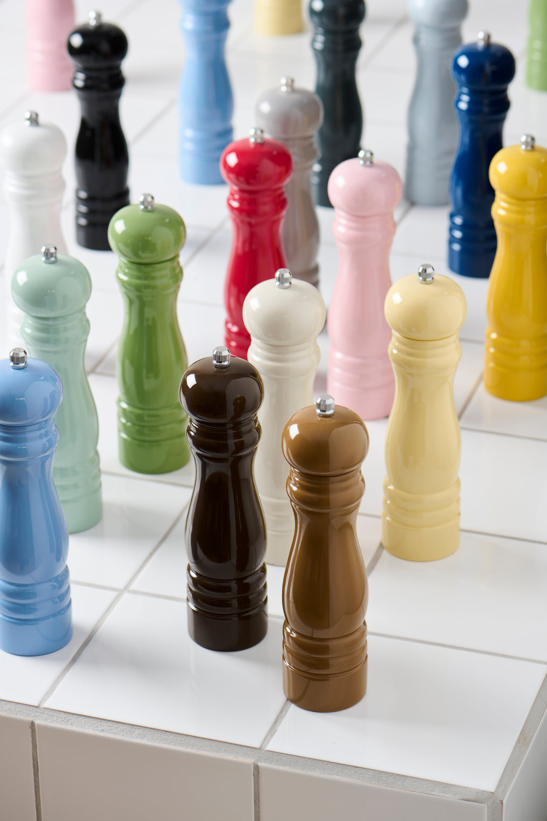 A collection of colorful pepper mills in various colors including blue, green, brown, pink, yellow, red, black, and white, arranged on a white tiled surface.