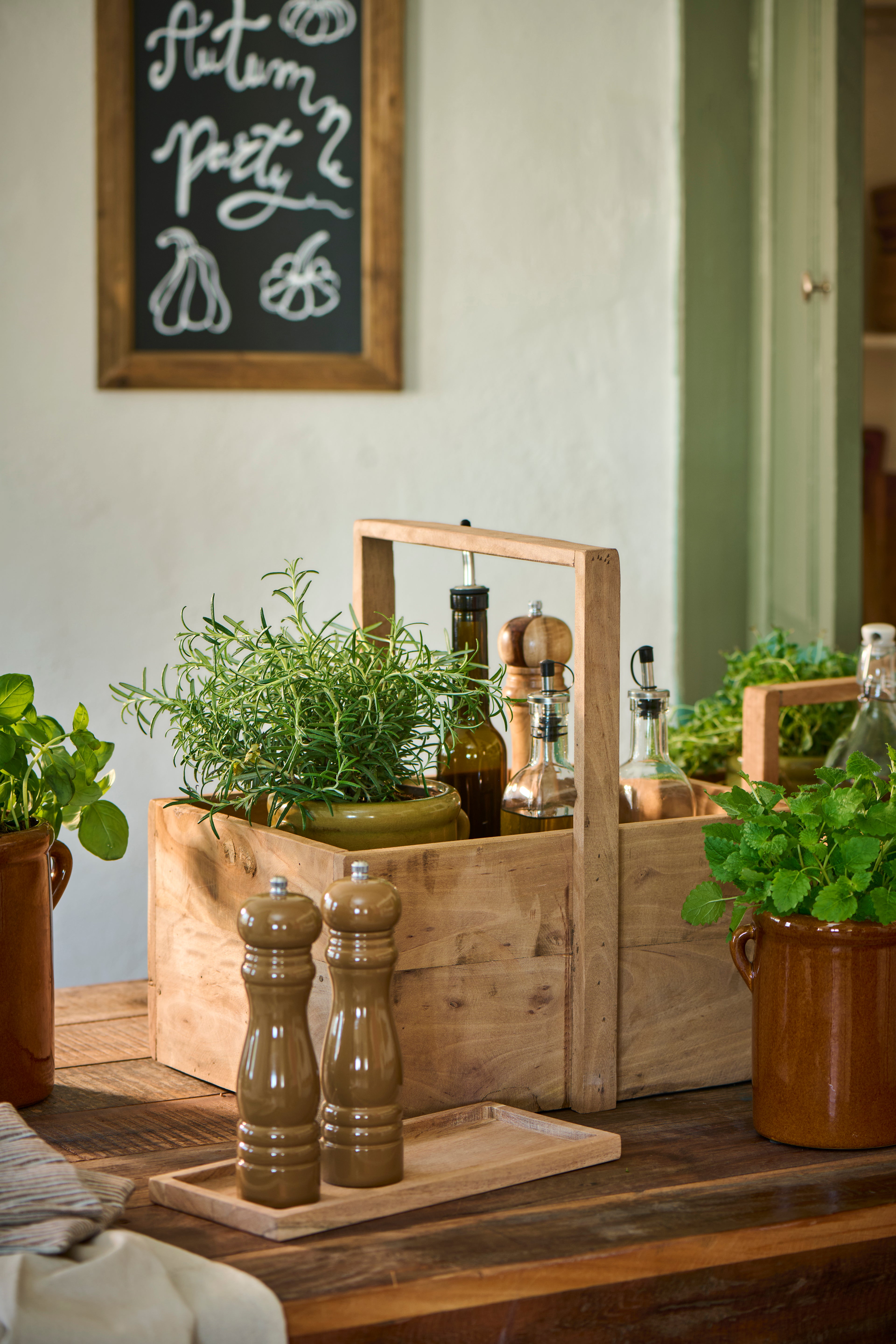 A rustic wooden herb carrier basket filled with fresh green herbs, set on a dark wood table alongside ceramic pots, glass bottles, and salt and pepper mills in a warm, styled kitchen setting.